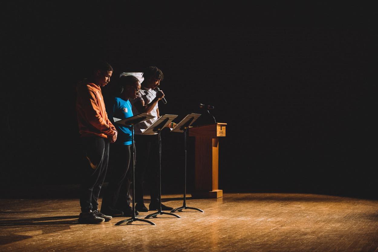 三个黑人学生, 穿便服, 站在黑人音乐台后面一排，读牧师的诗. Dr. 马丁·路德·金.'s 1963 "Letter from Birmingham Jail." The students are lit by a spotlight, 除了他们面前的木制舞台和他们右边的木制讲台之外，其他的都在他们面前, 在阴影. 他们正在参加烛光守夜活动，这是澳门威尼斯人网址马丁·路德·金周的活动之一.